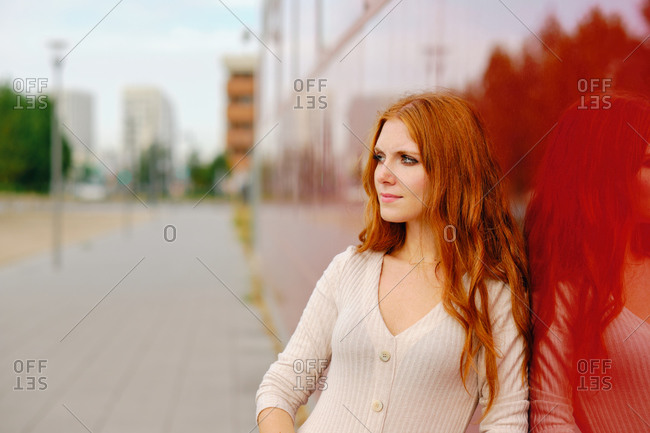 Young redhead female in elegant beige dress standing on pavement against bright red wall of modern urban building