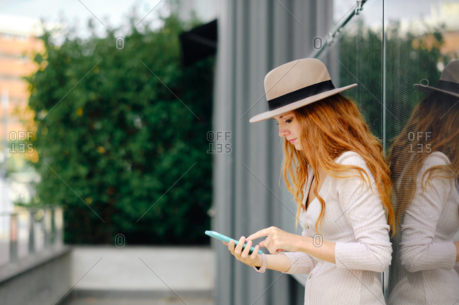 Young elegant redhead female in stylish outfit and hat browsing mobile phone while standing near glass wall of modern urban building