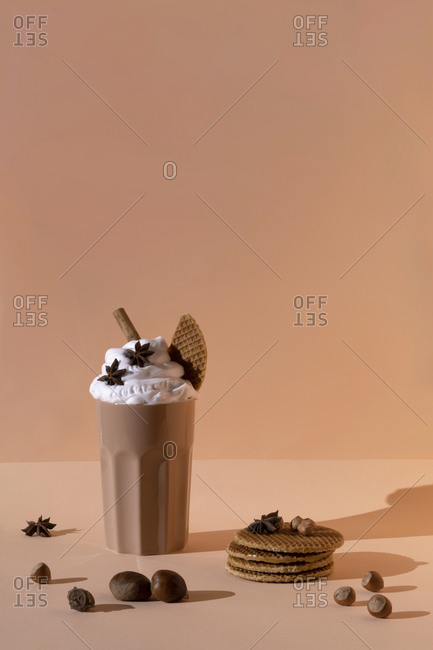 Glass with chocolate cocktail with whipped cream garnished with star anise and cinnamon stick placed on table with hazelnuts and cookies on pastel brown background
