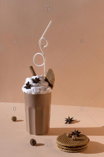 Glass with chocolate cocktail with whipped cream garnished with star anise and cinnamon stick placed on table with hazelnuts and cookies on pastel brown background