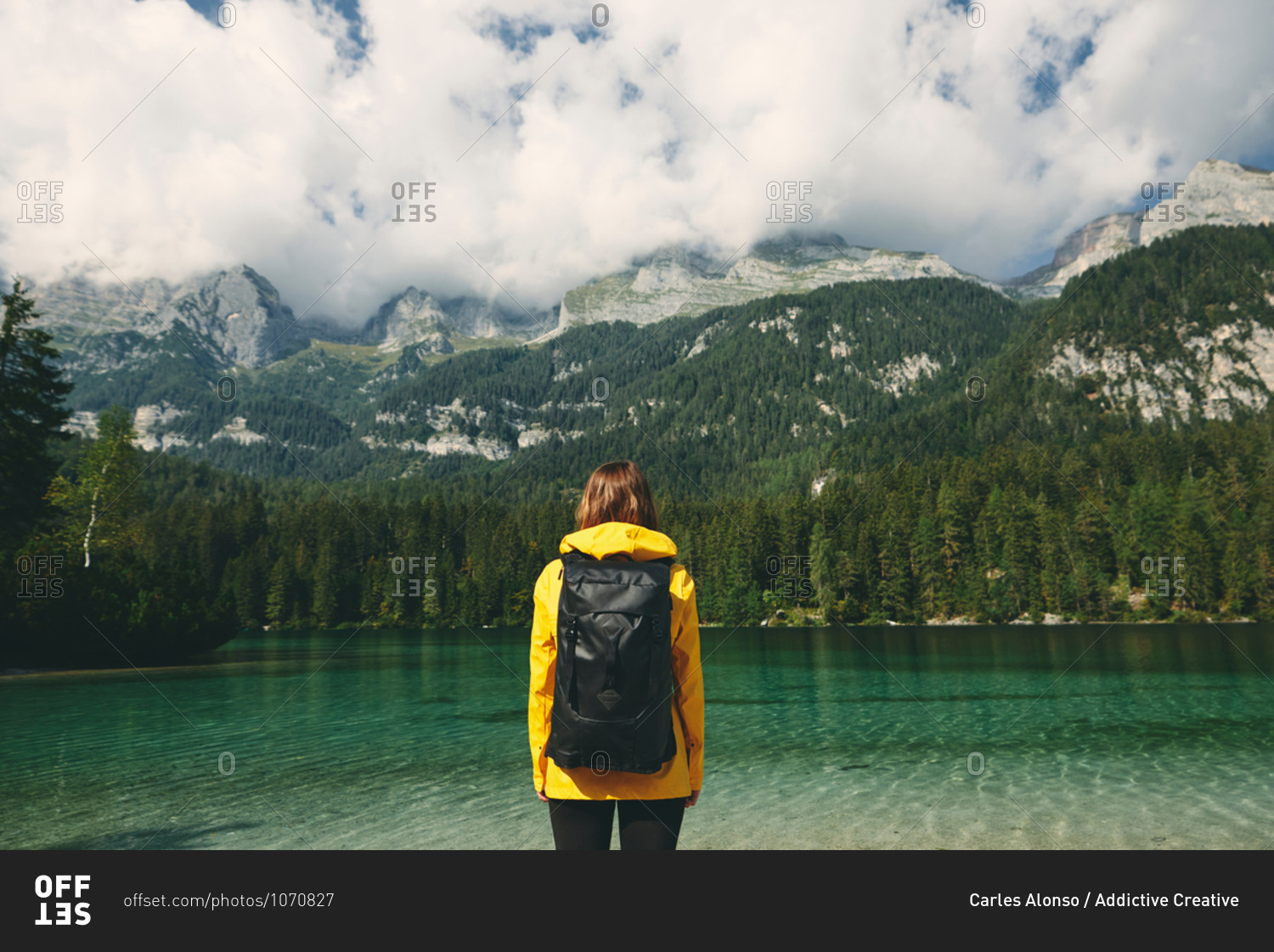 Back view of anonymous tourist with backpack admiring picturesque scenery of reflecting lake and mountains covered with forest in Italy
