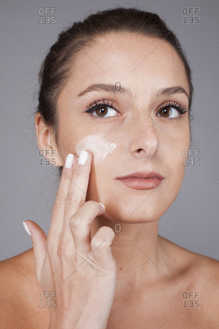 Young beautiful woman with healthy skin taking care of face with cream isolated on gray background looking away