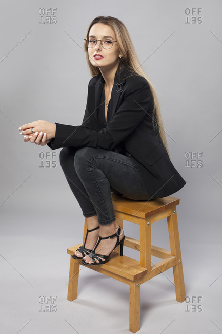 Side view of charming female model in smart casual style sitting on wooden stool on gray background and looking at camera