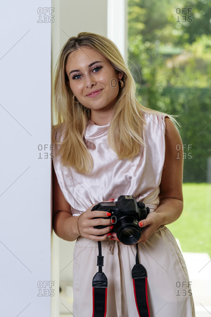 Cheerful professional female photographer standing leaning in wall with camera after working day
