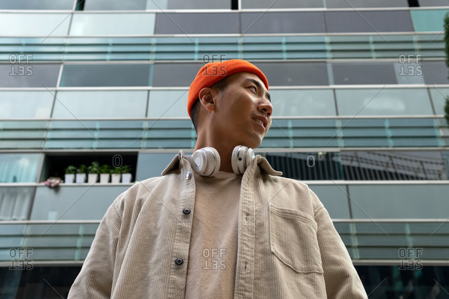 Low angle calm Asian male standing against contemporary condominium building and looking away in contemplation