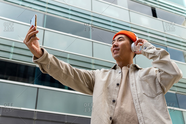 From below happy Asian male in casual wear and wireless headphones having video conversation through smartphone while standing against contemporary condominium facade