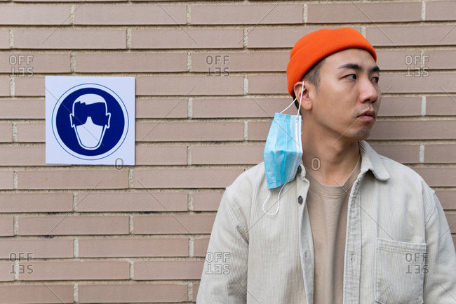 Serious Asian male braking rule by taking off mask standing near sign in building wall illustrating person in protective mask