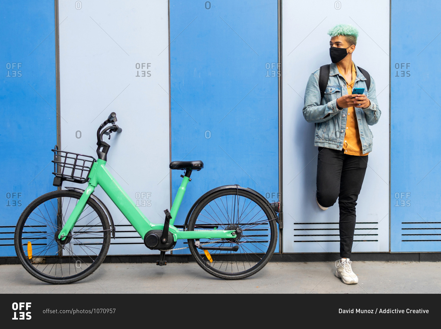 African American male hipster with dyed blue hair and in protective mask standing near wall with bicycle and browsing smartphone in city during coronavirus epidemic