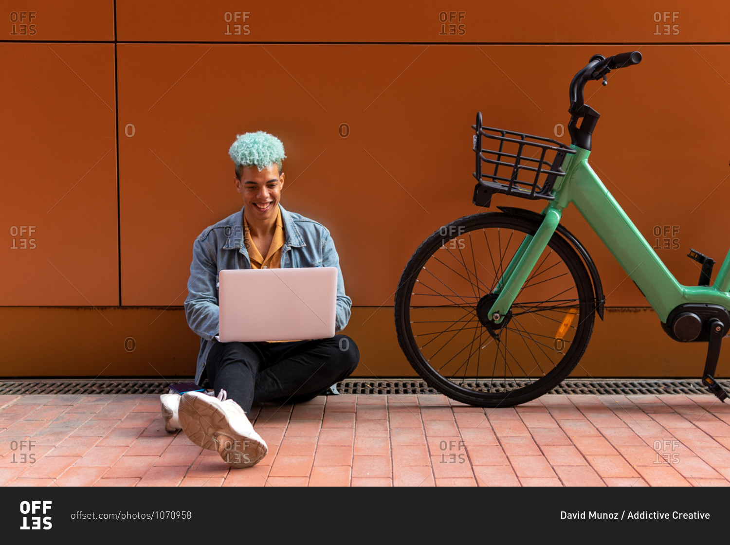 Smiling black male hipster with dyed hair using laptop while working on remote project on the street near modern bike