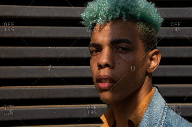 Dreamy black male hipster with blue hair standing on street and enjoying sunny day looking at camera