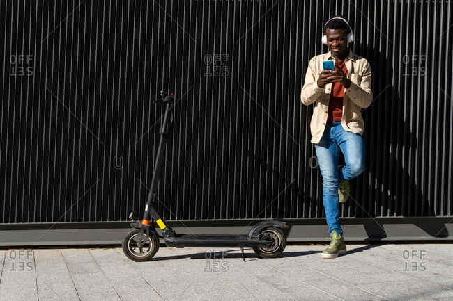 Happy African American male listening to music in wireless headphones while texting on mobile phone standing on metal wall with modern electric scooter along sunny street in city