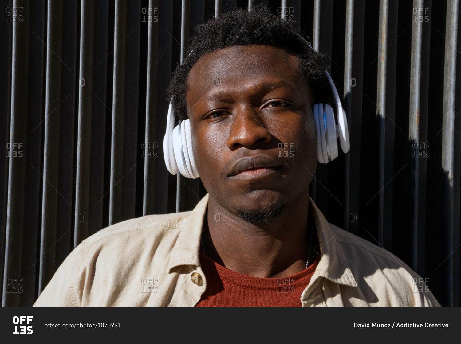 Tranquil African American male listening to music in wireless headphones while leaning on metal wall in street and looking at camera
