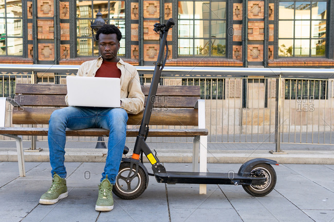 Concentrated African American male freelancer sitting on bench near electric scooter and working on project while using netbook in city