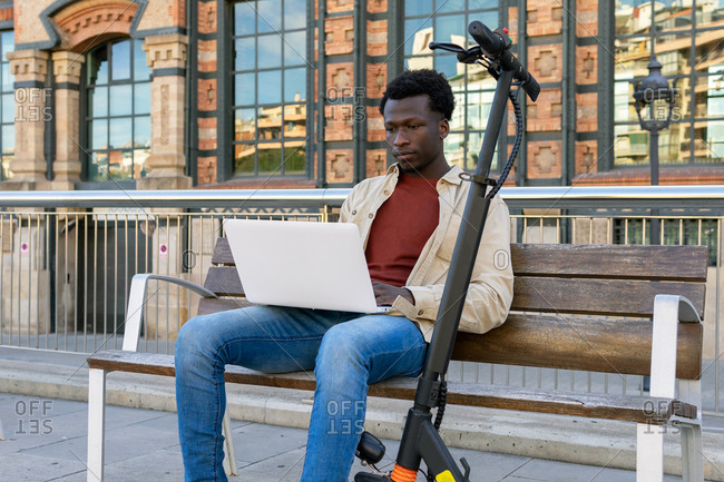 Concentrated African American male freelancer sitting on bench near electric scooter and working on project while using netbook in city