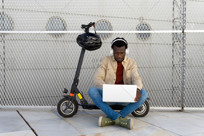 Black male sitting on parked electric scooter on street and messaging on social media via netbook while listening to music in headphones and enjoying weekend