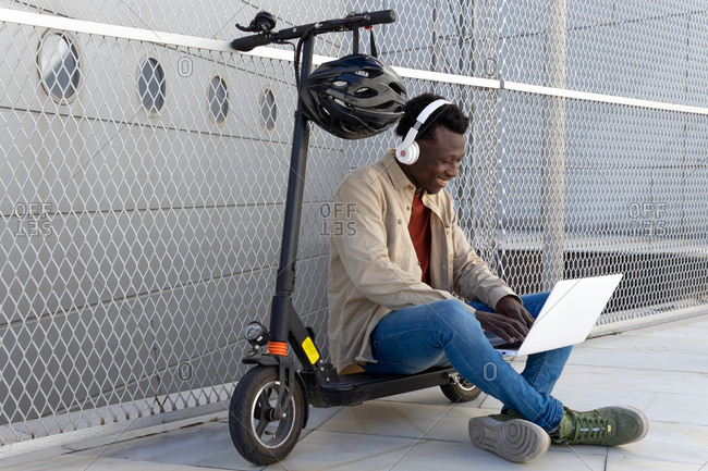 Cheerful black male sitting on parked electric scooter on street and messaging on social media via netbook while listening to music in headphones and enjoying weekend
