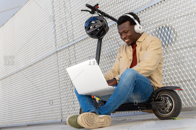 Low angle of cheerful black male sitting on parked electric scooter on street and messaging on social media via netbook while listening to music in headphones and enjoying weekend