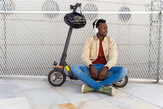 thoughtful black male sitting on parked electric scooter on street while listening to music in headphones and enjoying weekend looking away