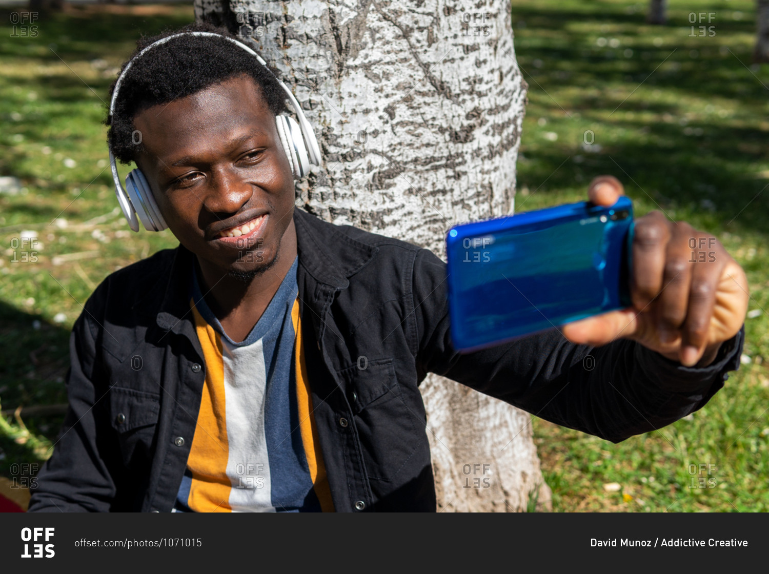 Handsome black male in wireless headphones sitting near tree in summer park and taking selfie on smartphone