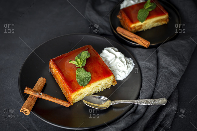Top view of black plate with portion of palatable gourmet apple pudding garnished with mint leaf and cinnamon sticks served with whipped cream on dark background