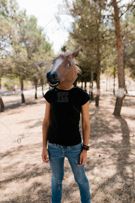 Unrecognizable male wearing weird rubber horse mask standing in park on sunny day
