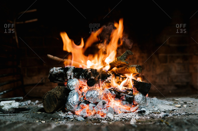 Potatoes in aluminum foil baked in burning flame of fire in cozy chimney in rustic house