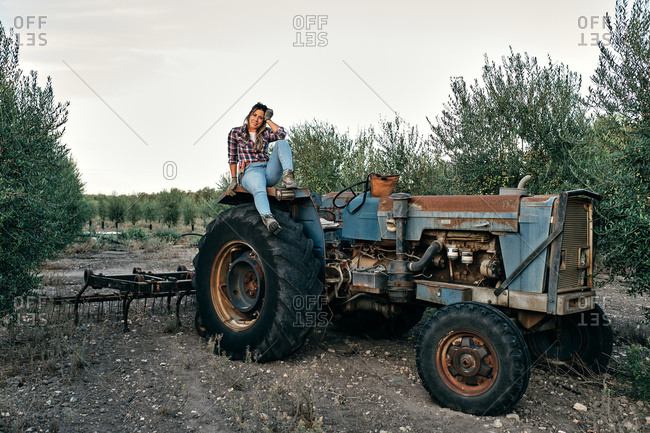 Satisfied adult female farmer sitting on wheel of agricultural machine during harvesting season on olive plantation