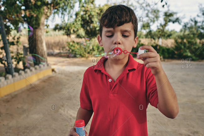 Adorable boy in summer outfit standing in park and blowing soap bubbles while having fun at weekend