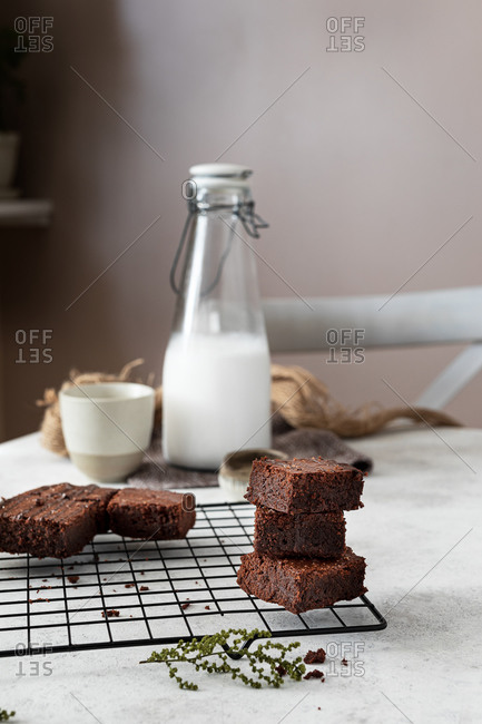 Pouring chocolate sauce from pot over stacked pieces of homemade brownie cake arranged on metal grid while preparing dessert in kitchen