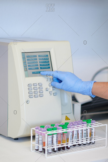 Crop unrecognizable medical technologist touching display of hematology analyzer near table with blood tubes in lab