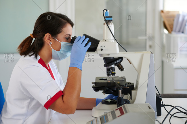 Side view of unrecognizable female medical specialist in uniform and mask working with microscope in laboratory