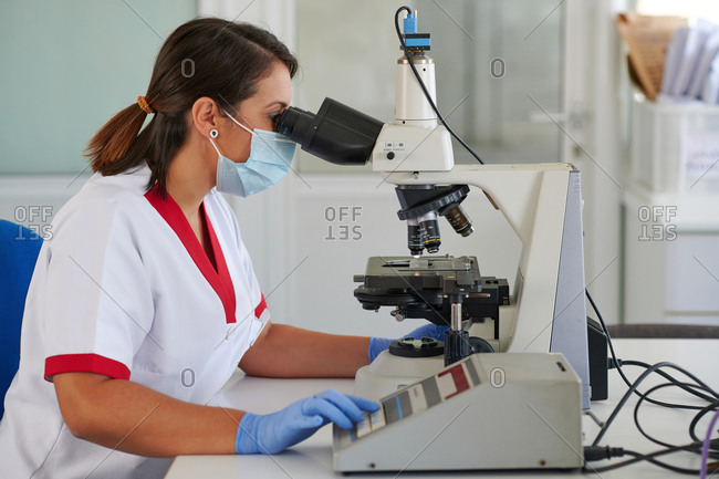 Side view of female medical specialist in uniform and mask working with microscope in laboratory