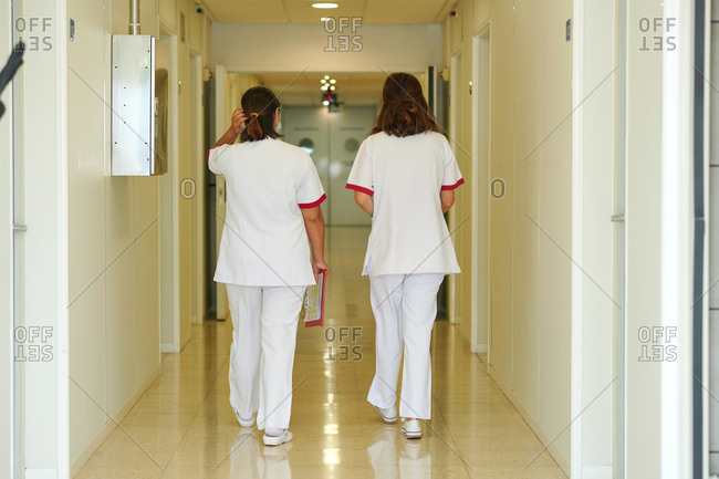Back view of unrecognizable female medical staff in white uniforms strolling on glowing passage floor in hospital
