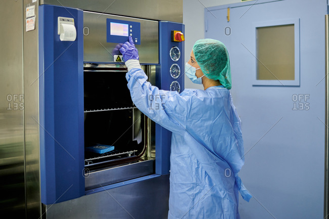 Side view female nurse wearing sterile protective uniform pushing buttons on modern sterilization machine while working in hospital