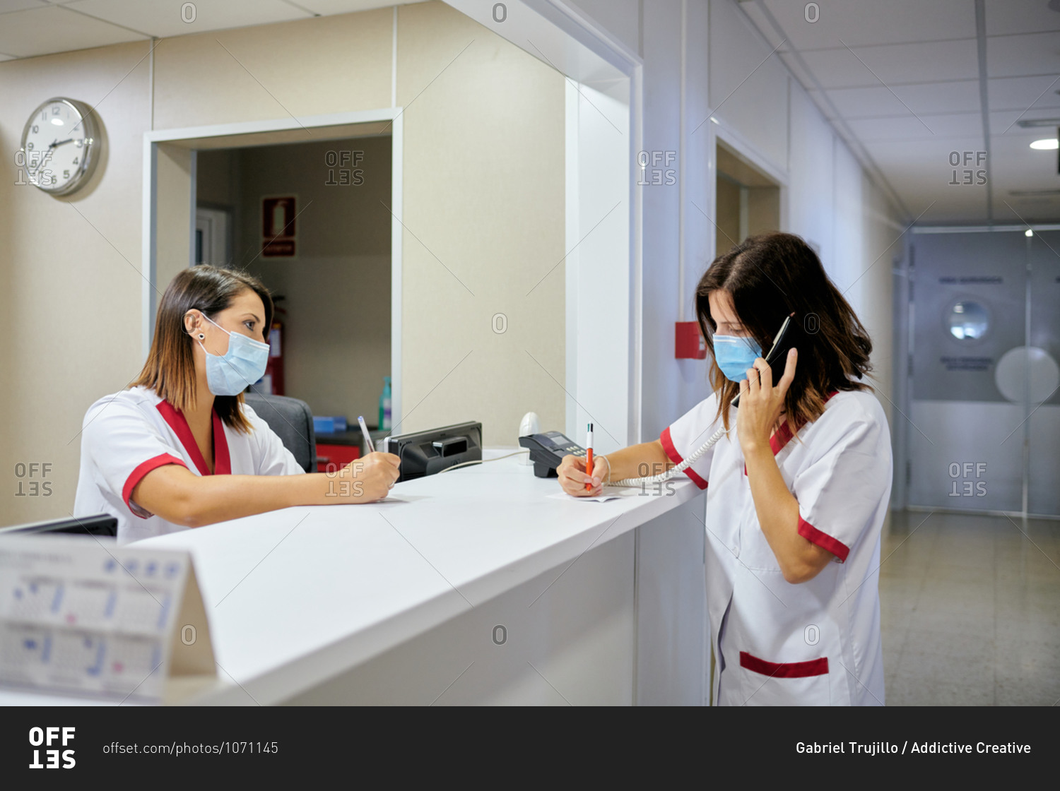 Concentrated female nurse wearing white medical suit and face mask taking notes in clinical record chart while standing near modern hospital reception