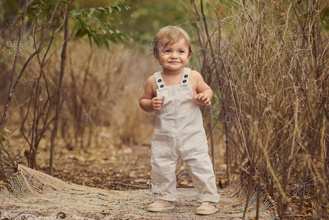 Full body of adorable toddler in overall standing near tall plants while smiling and looking away
