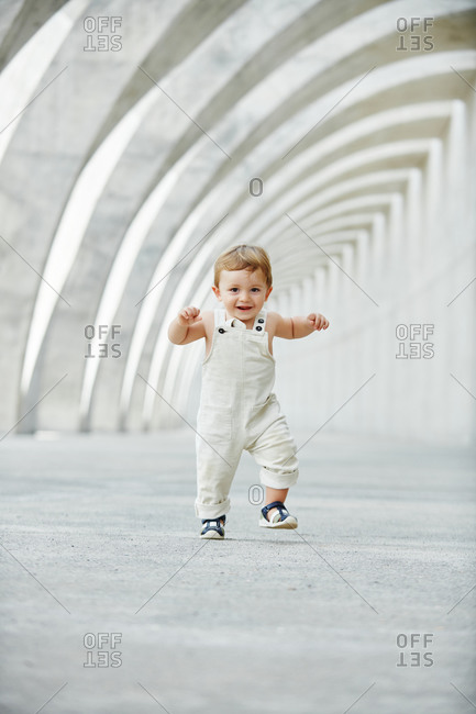 Delighted little kid in cute overalls walking along arched pathway in city and looking at camera