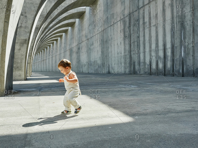 Delighted little kid in cute overalls walking along arched pathway in city and looking down