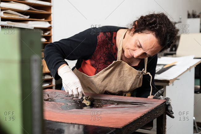 Mature female artist in apron and gloves covering engraved plate with ink while creating artwork in professional workroom