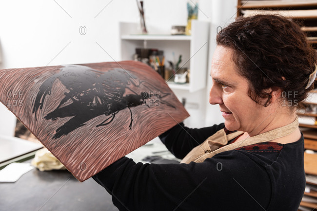 Side view of concentrated middle aged craftswoman holding piece of linoleum with inked pattern while making print in creative workplace