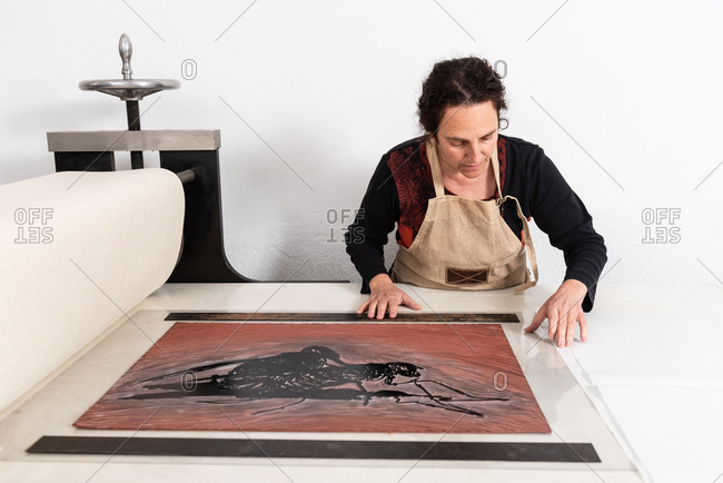 Focused middle aged female master putting sheet of linoleum with inked ornament on printing press while working in linocut technique in work studio