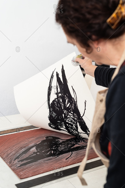 Side view of skilled female artist removing paper sheet with impression from linoleum piece placed on printing press machine while working in creative studio