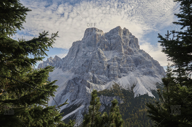 Picturesque scenery of high mountain in Dolomites under clouds near green coniferous forest under cloudy bright sky