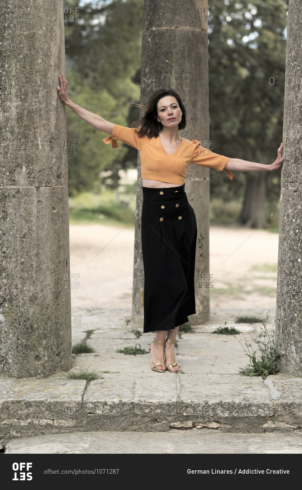Full body of thoughtful young slender female in crop top and long black skirt looking at camera while standing between tall thick columns in park in daytime