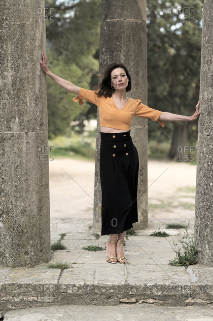 Full body of thoughtful young slender female in crop top and long black skirt looking at camera while standing between tall thick columns in park in daytime