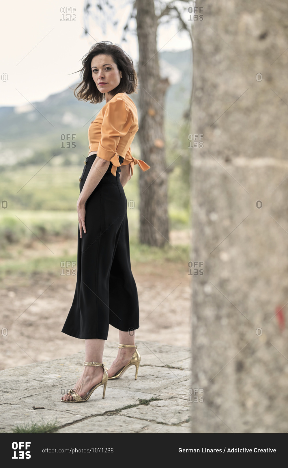Side view full body of thoughtful young slender female in crop top and long black skirt looking at camera while standing between tall thick columns in park in daytime