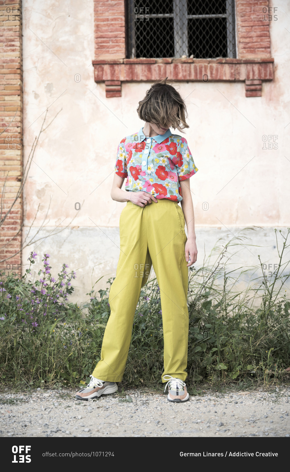 Full body of faceless female in shirt with floral print and light green trousers looking down while standing under window of old construction in daytime