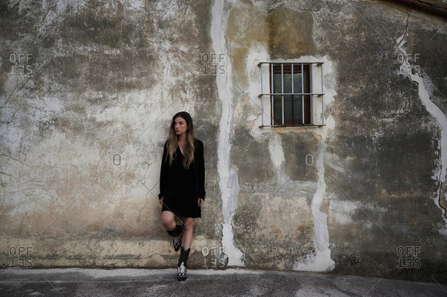 Full body of young female in casual black dress and boots leaning back against shabby stone wall of aged building with small window