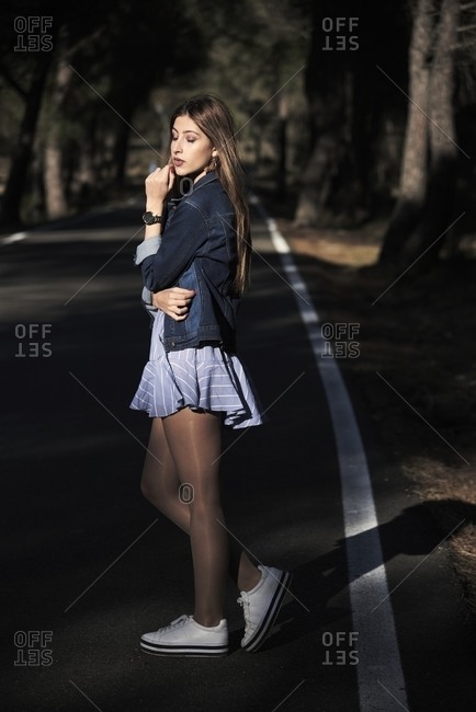 Side view of peaceful female in hipster clothes standing on empty asphalt road and enjoying sun with closed eyes