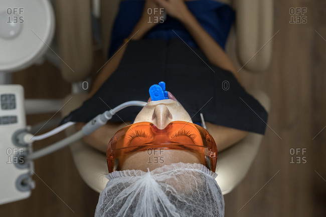 From above female patient in laser safety goggles with dental expander and saliva ejector in mouth getting treatment procedure in modern clinic